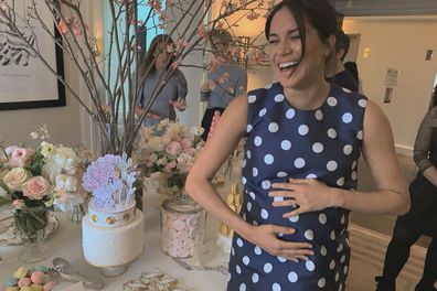 Candid of Meghan Markle at her  2019 baby shower in New York laughing and holding baby bump beside a cake. Meghan Markle is wearing a navy dress with white polka dots.