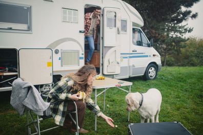 A woman is sitting on a chair in front of a camper, eating a banana and giving some banana to her dog