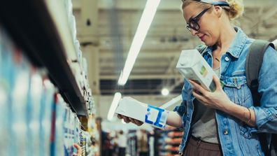 Beautiful blonde women wearing glasses, in a denim jacket and a skirt, shopping for milk in the supermarket. She is holding two cartons of milk and choosing which one to buy.