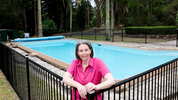 Sandra stands inside the fence of her pool. She's wearing a pink shirt. The pool is a clear blue.