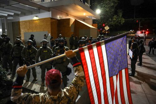 A man holds an American flag as law enforcement officers guard the enterance to ICE headquarters in south Portland, Oregon.