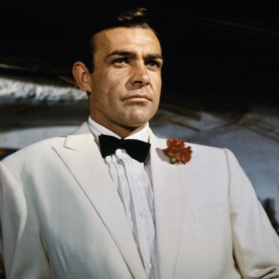 Waist-up portrait of Sean Connery, as James Bond, leaning against a bar and looking out across the room. Connery is wearing a white tuxedo and bow tie with a red carnation in his lapel. CONNERY; SEAN BOND; JAMES. 99/99/1966 35 U CT4X5