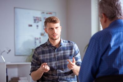 Confused young man gesturing while discussing with therapist. Male mental health professional is with university student in meeting. They are talking at lecture hall.