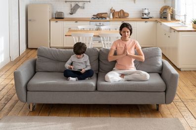 Young fit mother doing yoga exercise, sitting in lotus pose on couch together with little son at home. Calm child using gadget, playing a game on tablet, sit on sofa. Stress relieve, wellness concept.