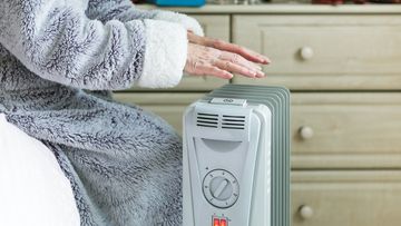 A retired senior woman in her 70s sits at home inside her cold house in winter. It is so cold that she is wrapped up in warm winter clothing, and is holding her hands over an electric heater for some extra warmth and comfort. Selective focus with room for copy space.