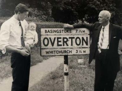 Peter Overton as a baby, being held by his father, as his grandfather looks on at the sign for the village of Overton in England.