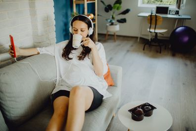 Young Caucasian pregnant woman with headphones listening to music at home while relaxing on the sofa, drinking coffee/tea, and using a smartphone.