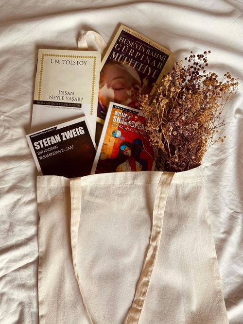 Stock image of novels and dried flowers emerging from the top of a canvas tote bag, which appears to be laying on a bed.