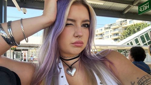 Uma jovem de cabelo roxo pisca para a câmera enquanto tira uma selfie. Ela está usando joias de prata e parece estar sentada em uma estação de trem.