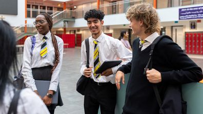 A eye-level medium close-up of a group of teenage students who are waiting to go into class in the high school they go to in the North East of England. They are carrying their books and bags and laptops and devices.