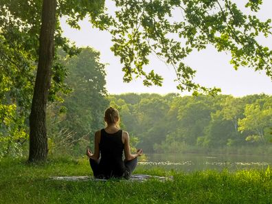 Back view of unrecognizable woman practicing yoga in summer outdoors. Rear view of female sitting in meditation pose on yoga mat in lotus position, raising hands up, doing stretching exercise outside