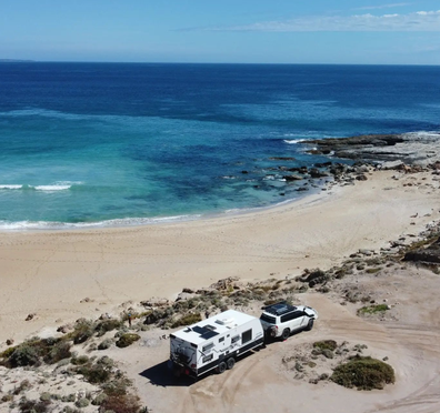 A car with attached caravan is parked on the sand next to a stunning blue beach. Auswide Explorers Instagram