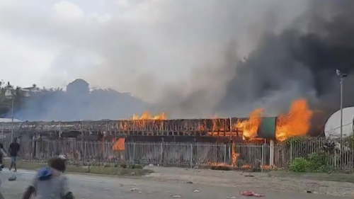 Des flammes sauvages déchirent une propriété alors que des personnes, potentiellement des manifestants, s'attardent à proximité.