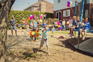 Little girl hitting pinata at birthday party in suburb backyard. Lots of colorful ballons in the background with family and guests. Some kids are sitting on the trampoline watching with interest. This was taken at a real birthday party organized on a hot and sunny spring day. Horizontal.