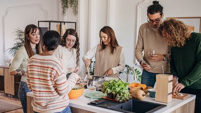 Group of people, diverse male and female friends preparing a meal and drinking wine together in domestic kitchen,