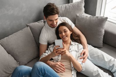 Caucasian couple man and woman in casual clothing resting in living room at home and looking at smartphone