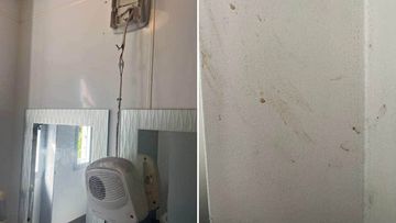 A bathroom heater hangs by an electrical wire, while a wall is smeared with unknown matter.