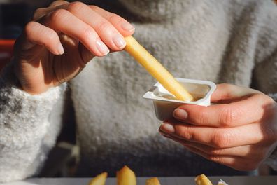 A woman holds French fries in her hand, eating them in a cafe or restaurant. A girl in a white knit sweater dips a slice of potato in garlic sauce. The concept of fast food, snacking, junk food.