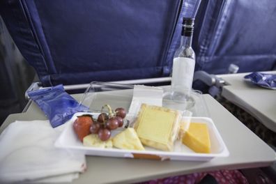 Food and drink that has been purchased on a plane is on top of the fold down tray of an airplane. It contains cheese, fruit and crackers. There is an empty bottle of wine on the tray. There are no people in the picture that was shot with a Canon 5D Mark 3 camera.