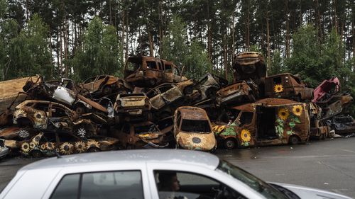 Vehicles destroyed by Russian attacks are piled up in a lot as a driving instructor, foreground, talks to his student driver in Irpin, Ukraine.
