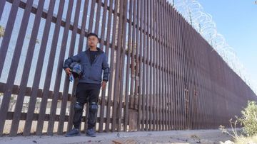 Wang Qun stands before the high steel fence on the US-Mexico border.