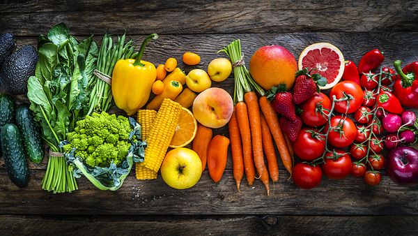 Top view of a large assortment of healthy fresh rainbow coloured organic fruits and vegetables arranged side by side on rustic wooden table. The composition includes carrots, onion, tomatoes, avocado, corn, green bean, cucumber, broccoli, spinach, apples, strawberries, mango, grape fruit, peach, oranges, bell pepper, radish among others. 