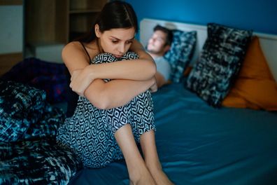 Sad girlfriend sitting on bed edge, holding her legs after fight, her partner is lying in bed in background, his face is lit by smart phone