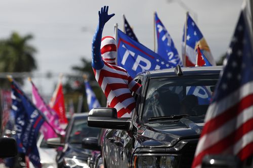 A man wearing a flag-themed body stocking waves from a car as hundreds of vehicles gather at Tropical Park
