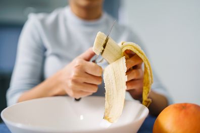 Caucasian woman sitting by the table at home cutting banana - Adult girl female preparing fruit salad at home - healthy eating concept copy space close up