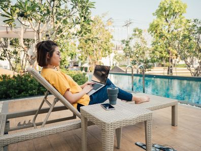 Freelancer works with laptop and smartphone and lies on a sun lounger by the pool in the resort on summer vacation.