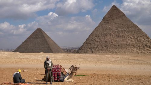 A picture taken on December 17, 2021 shows men resting next to a camel with the Great Pyramid of Khufu (Cheops) (L) and Pyramid of Khafre (Chephren) (R) in the background at the Giza pyramids
