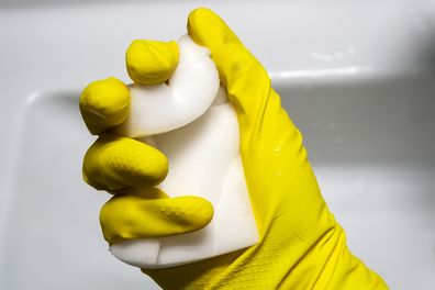 A hand in a yellow rubber glove squeezes a wet white melamine sponge against a white bathroom. 