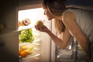 Close up image of a young woman, having a midnight snack - eating an apple, in front of the refrigerator.