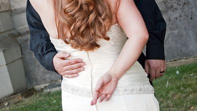 A bride stands with her back toward the camera, with her fingers crossed.