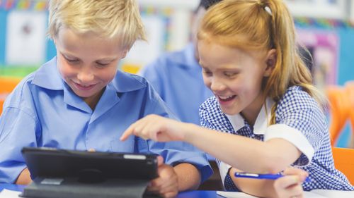 Students using a digital tablet in a classroom. There is a boy and a girl, both are wearing school uniforms and are happy and smiling.