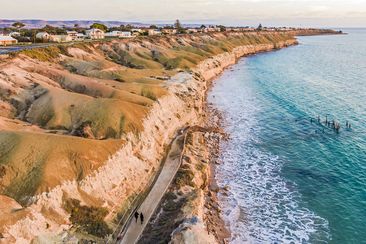 Aerial view Port Willunga sunset, clifftop houses with views of St Vincent Gulf, cliffs, rock face, ocean, unidentifiable couple in black walking on footpath, old jetty ruin and rolling hills in the distance.