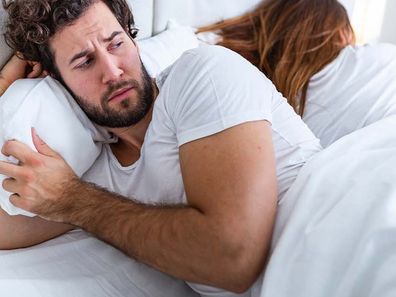 Man and woman lying in bed. The man is in front and looking suspicious of the wife, who is turned the other way, asleep.