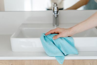 Close-up view of cleaning the white washbasin with a blue microfiber cloth. Woman hand removing drops and dirt in the modern bathroom. Concept of housekeeping and housework