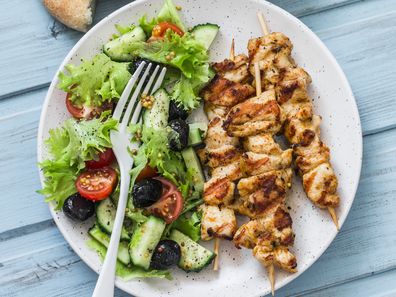 Chicken souvlaki kebabs, sun dried olives, tomatoes, cucumbers green salad and focaccia on blue background, top view