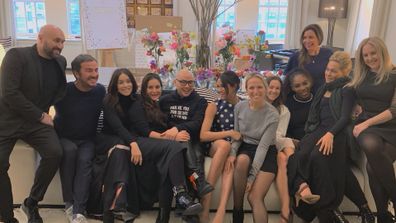 Meghan Markle surrounded by friends at her baby shower in 2019 in New York. Twelve people huddled on a couch for group photo, some smiling at camera, others looking in the distance. Most wearing black, one woman in grey and Meghan Markle in navy dress with white polka dots.