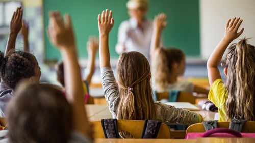 Rear view of large group of students raising their arms to answer the question on a class at elementary school.