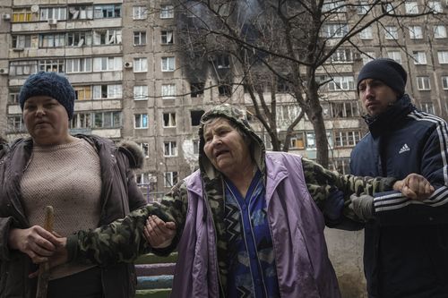 Des personnes aident une femme âgée à marcher dans une rue avec un immeuble touché par des bombardements en arrière-plan à Marioupol, en Ukraine, le lundi 7 mars 2022.