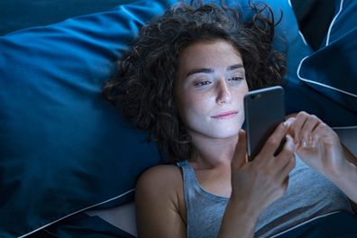 Top view of young woman using a smartphone while lying on bed in the night. High angle view of pretty girl messaging on smart phone before sleeping at night. Addicted woman suffering from insomnia and chatting and surfing on the internet with her cellphone late in night.
