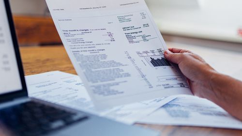 Uma pessoa analisa sua conta de energia enquanto usa um laptop em casa. O documento inclui vários encargos e informações sobre o uso de eletricidade.