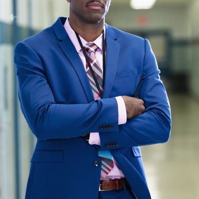 A mature African-American man standing in a school hallway, looking at the camera with his arms crossed. He is a teacher or principal.