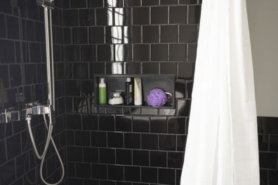 A shot of a black-tiled bathroom with bathroom toiletries on a marble shelf and a shower faucet and a curtain.