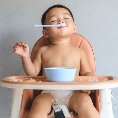 Happy infant Asian baby boy eating food by himself on baby high chair and making mess with copy space.