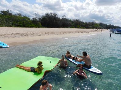 Group at Shelly Horton Christmas party swimming on a beach. One man sitting on long board with paddle, another woman lying on floating mat.