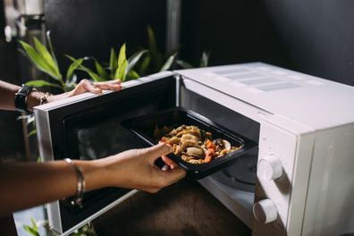 A side view of an unrecognizable woman standing in the kitchen and opening the microwave while holding a plastic container of food.