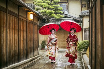 Full length of young maikos holding red umbrellas during rainy season. Beautiful geisha girls wearing traditional dress called kimono. They are walking on wet street.